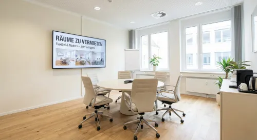 Ein heller Konferenzraum mit einem großen Bildschirm an der Wand, auf dem "RÄUME ZU VERMIETEN" steht, umgeben von einem runden Tisch mit Stühlen.